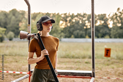 Portrait of good-looking fit woman Hunter with gun rifle, posing at camera. Caucasian young lady in cap, protective headphones and eyeglasses is looking confident and serious, ready to shoot