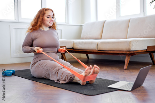 fat obese woman exercising with rubber bands according to online sports video on laptop at home. Plus size caucasian redhead lady doing domestic workout in front of modern gadget, in living room
