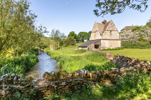 The old stone dovecote (c.1600 AD) beside the infant River Windrush as it flows through the Cotswold village of Naunton, Gloucestershire UK