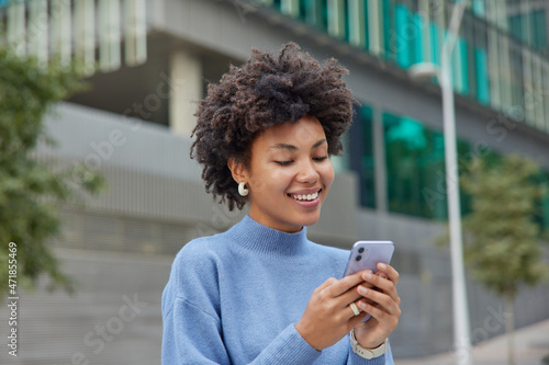 Half length shot of positive glad woman in blue turtleneck poses in urbanity with mobile phone explores new city downloads application has chat conversation. People technology and free time.