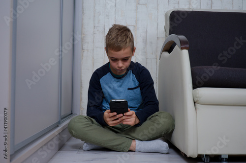 młody chłopak siedzi na podłodze między fotelem a szafą używa telefonu 