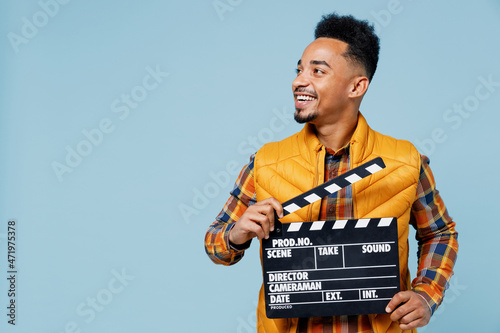 Jubilant exultant young black man 20s years old wear yellow waistcoat shirt looking aside holding classic black film making clapperboard isolated on plain pastel light blue background studio portrait
