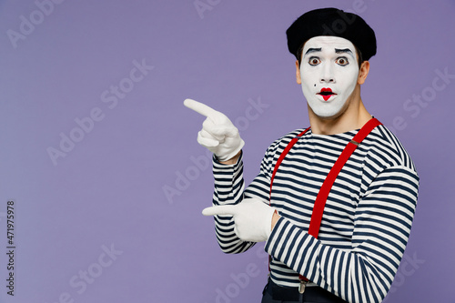 Surprised ecstatic young mime man with white face mask wears striped shirt beret pointing aside on workspace area copy space mock up isolated on plain pastel light violet background studio portrait.