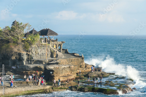 Crowds visiting the Tanah Lot Temple, Bali, Indonesia, located on the cliffs by the seashore. The waves are splashing on the cliffs and smaller rocks. Clear and sunny day. Power of the nature