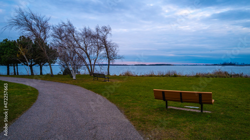 The curving footpath and an empty bench in the park with bare trees. Moody cloudscape at dusk. Twilight seascape over the green at Nut island Pier Park in Quincy, Massachusetts.