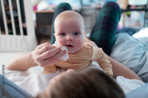 Young fatherwipes drool with white paper handkerchief of his newborn baby girl in bed.