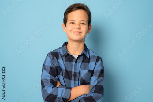 Smiling boy wearing plaid shirt with arms folded standing over blue background.