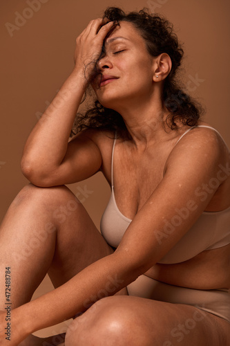 Pretty woman with imperfect body, cellulite and stretch marks wearing beige underwear hiding half of her face posing against colored background with copy space