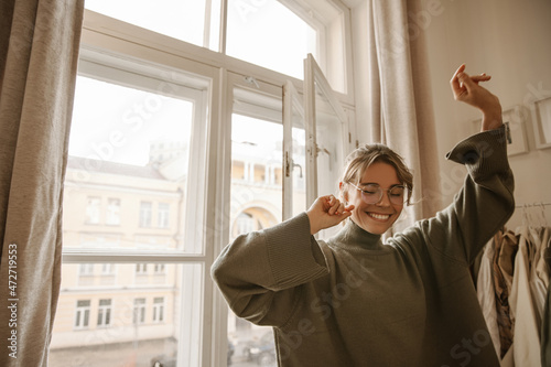 Funny young fair-skinned woman dancing with her eyes closed alone at home in daytime. Blonde with hair pulled back wears translucent glasses and oversized sweater. Freedom and happy lifestyle concept