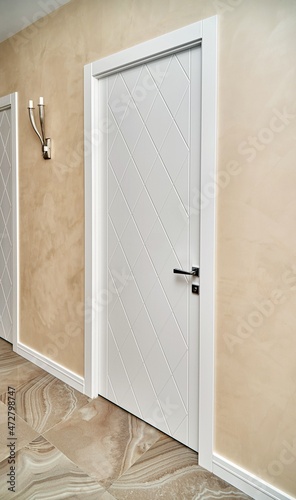 Closed contemporary white door decorated with rhombus pattern in corridor with chandelier on beige wall in restored building