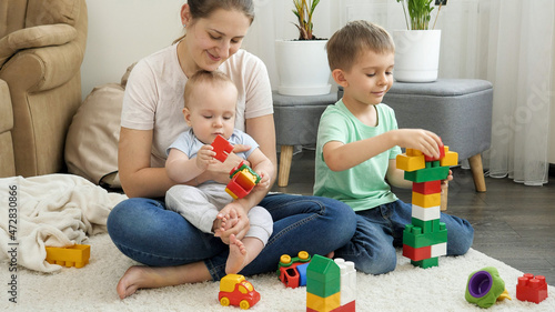 Happy children with mother playing on carpet and building toy tower. Concept of family having time together and children development