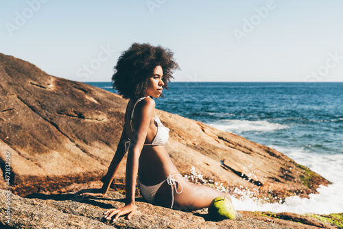 A ravishing young African American woman in white bikini and with curly afro hair is enjoying a sunset on the beach of a tropical resort while sitting on a rock in front of the ocean