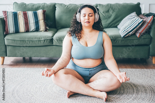 Curly haired plus size young woman in wireless headphones turns on music and meditates sitting in lotus position on floor mat against green sofa