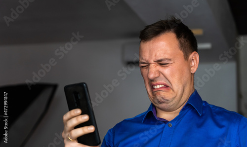 Annoyed millennial man with disgust look at smartphone screen indoors. Portrait of manager cringe and looking at phone screen with squeamishness, bad joke or inappropriate content