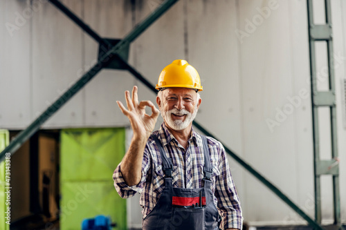 A happy senior blue-collar employee with a protective helmet is standing in the factory and showing okay gestures. A worker is approving, business is fine. A worker in a factory.