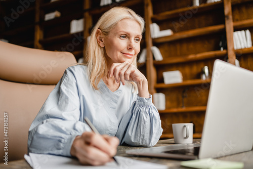 Smart caucasian mature middle-aged businesswoman CEO boss leader teacher writing, taking notes on laptop, e-learning , working remotely online in home office