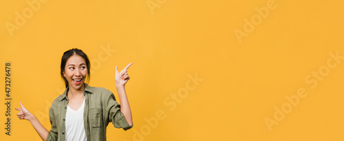 Portrait of young Asian lady smiling with cheerful expression, shows something amazing at blank space in casual clothing and standing isolated over yellow background. Panoramic banner with copy space.