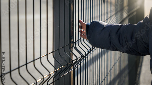 Closeup of little boy walking by the metal fence and touching it with hand. Concept of poverty, immigration, bullying and kids stress