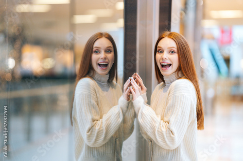 Portrait of excited cheerful female shopaholic standing by showcase and saying wow looking at camera, blurred background. Happy young woman looking at fashion display in shop window.