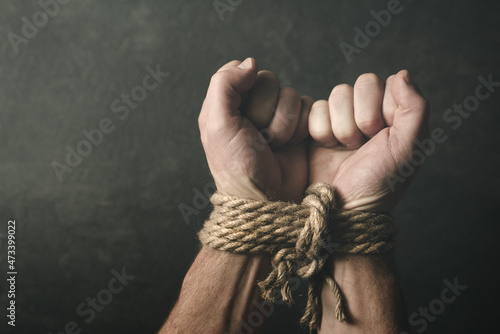 Male hands tied with a rough rope at the wrists close-up on a dark background, soft focus. Conceptual image of violence, slavery, dependence, lawlessness, lack of freedom