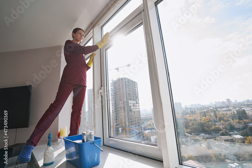 Pensive housecleaner in rubber gloves wiping window-frame