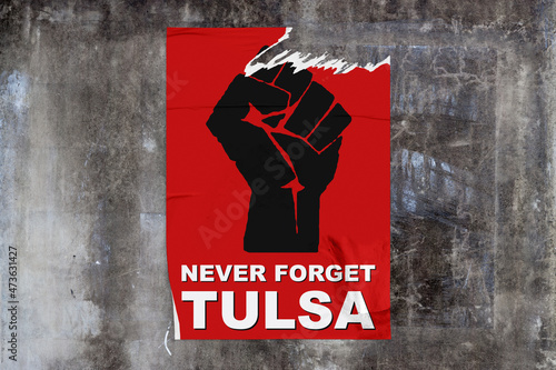 Full-frame weathered concrete wall with a torn red poster in the middle dispicting a black fist with "Never forget Tulsa" written bellow.