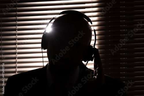 An FBI secret agent listens with headphones and records a conversation against the background of a window with blinds, silhouette lighting, selective focus, dark tone.