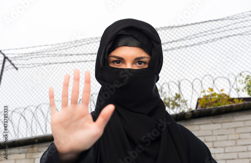 migration crisis, freedom and security concept - woman in hijab showing stop sign over barbed wire fence background