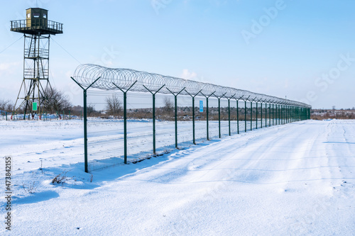 Border fence with barbed wire in front of the surveillance tower in a snow-covered field against a blue sky. Border protection and counteraction to illegal migration concept