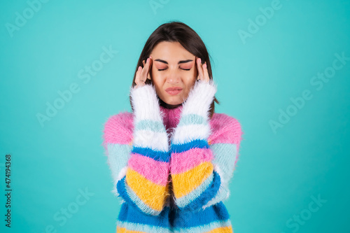 Young woman in a bright multi-colored sweater on a blue background, suffering from a headache, pressing her fingers to her temples