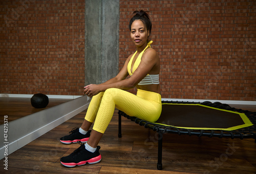 African woman with slim body and flat belly in bright yellow tracksuit sitting on a trampoline in lotus pose and smiles looking at camera. Fitness, sport, meditation, yoga, active lifestyle concept