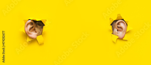 Two woman's ear through a torn holes in yellow paper. The concept of eavesdropping, espionage, gossip, tabloids and the yellow press. Background with copy space.