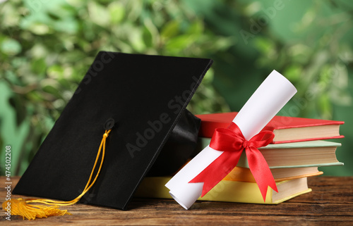 Graduation hat, books and diploma on wooden table against blurred background