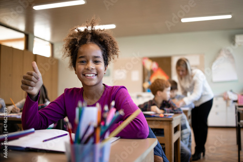 Portrait of smiling Afro American school child sitting in classroom with classmates and teacher in background.