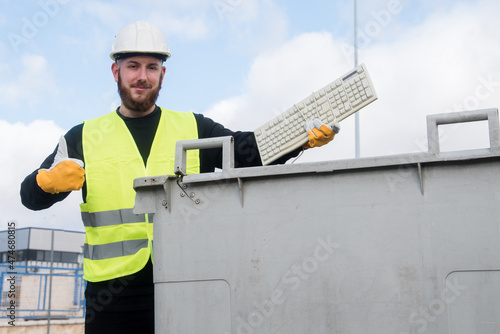 Trabajador del centro de reciclaje arrojando un viejo teclado de computadora a un contenedor de basura. Hombre con casco y guantes protectores y un chaleco reflectante