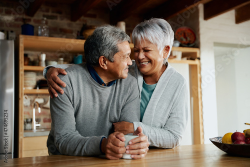 Happily retired elderly biracial couple smiling at each other. Wife holding husband in modern kitchen. 