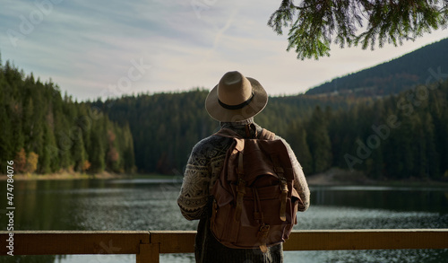 Rear view traveler man wearing hat with backpack standing by mountain lake with calm blue water, enjoying amazing landscape with forest and mountains