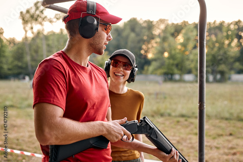 Handsome male shooting instructor teaches the backslider how to handle weapon on training course in outdoor range. cheerful teacher and woman in protective spectacles and headset have fun, laugh
