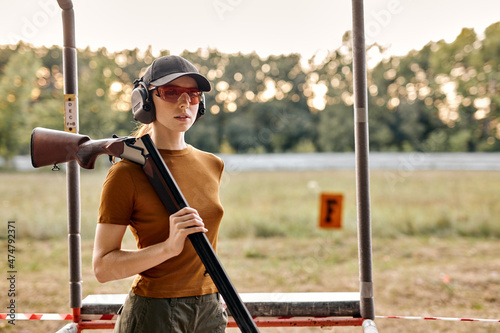 Good-looking caucasian lady with firing gun in outdoor academy shooting range, field in the background. Young beautiful woman in cap, headset and spectacles posing at camera after successful training