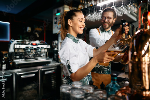 Female bartender pours beer draft beer while working with colleague in bar.