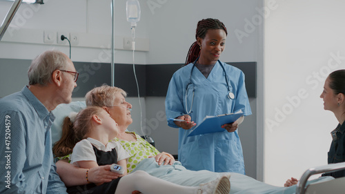 Close up of medical assistant explaining disease to patient and family in visit in hospital ward. Woman nurse talking about treatment and recovery to visitors and retired person with sickness.