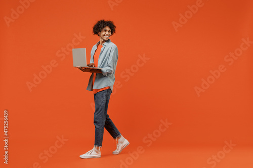 Full body young student black man 50s in blue shirt t-shirt hold use work on laptop pc computer look aside on workspace area mock up isolated on plain orange color background People lifestyle concept