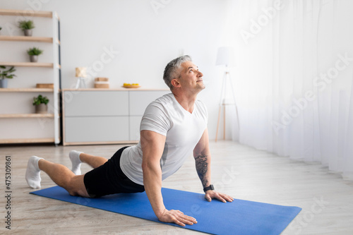 Active senior man doing cobra pose on yoga mat, exercising in living room interior, empty space