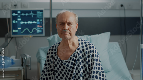 Portrait of elder patient with nasal oxygen tube standing on bed in hospital ward at medical clinic. Sick old man with oximeter connected at heart rate monitor to check progress