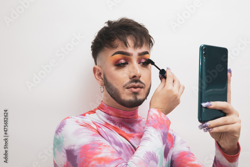 A young man is putting on makeup while using his mobile as a mirror on a white background with fashionable clothes.