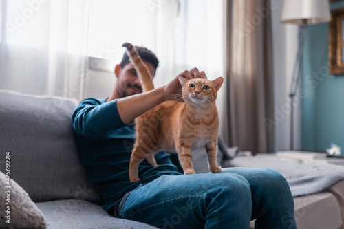 young man sitting on a gray sofa caresses the head of a brown tabby cat