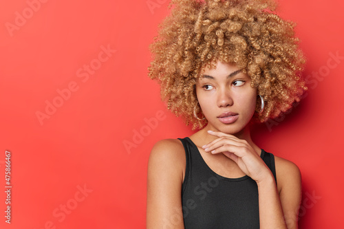 Pensive curly haired woman with natural beauty touches jawline concentrated aside dressed in black t shirt being deep in thoughts isolated over red background copy space for your promotional content.