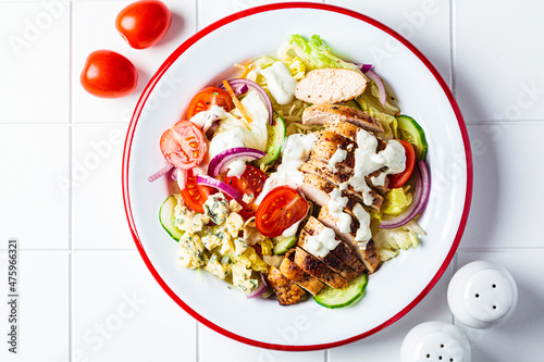 Buffalo chicken salad with blue cheese, tomato, cucumber and ranch dressing, white tile background.
