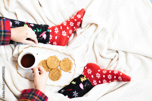 Closeup shot of a person in cozy Christmas pajamas and socks drinking tea and eating cookies in bed