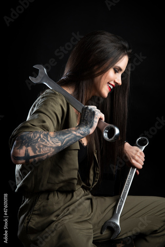 Atrakcyjna uśmiechnięta dziewczyna - mechanik z tatuażami, pozująca z dwoma bardzo dużymi kluczami. Zdjęcie studyjne na czarnym tle, oświetlenie z 2 lamp.